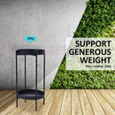 nzmegabuy 2-tier black metal plant stand on a wooden floor against a concrete and green leaf wall background
